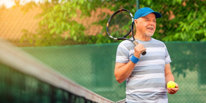 Senior man smiling on tennis court holding racket and ball