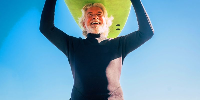 Smiling senior man holding surfboard overhead on beach