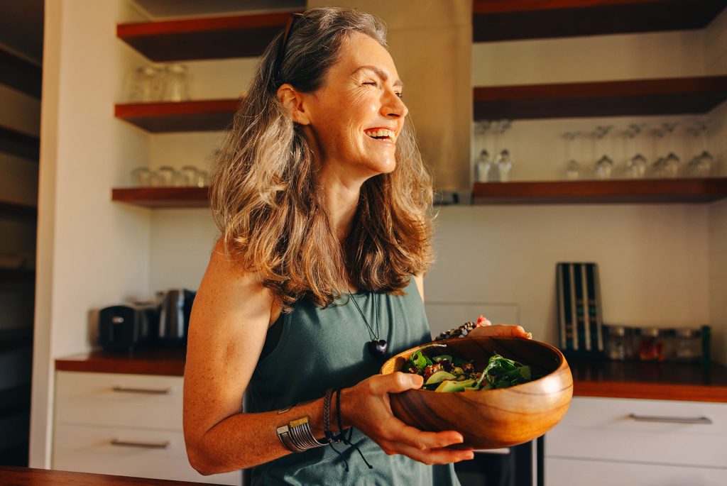 Smiling mature woman holding a large wooden bowl of fresh green salad in her kitchen
