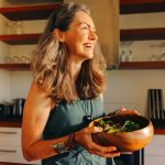 Smiling mature woman holding a large wooden bowl of fresh green salad in her kitchen
