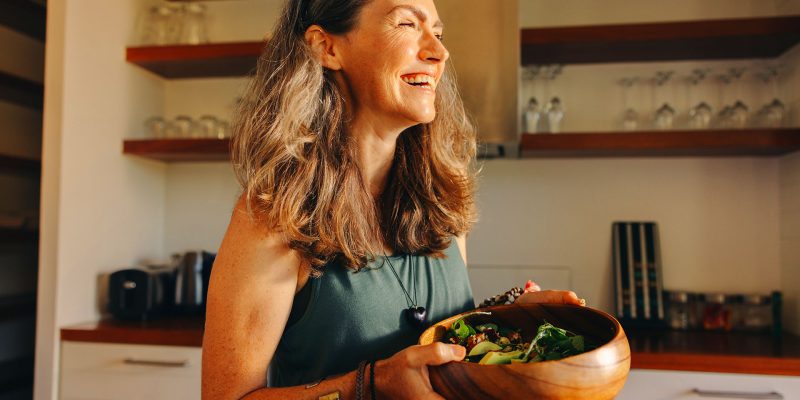 Smiling mature woman holding a large wooden bowl of fresh green salad in her kitchen